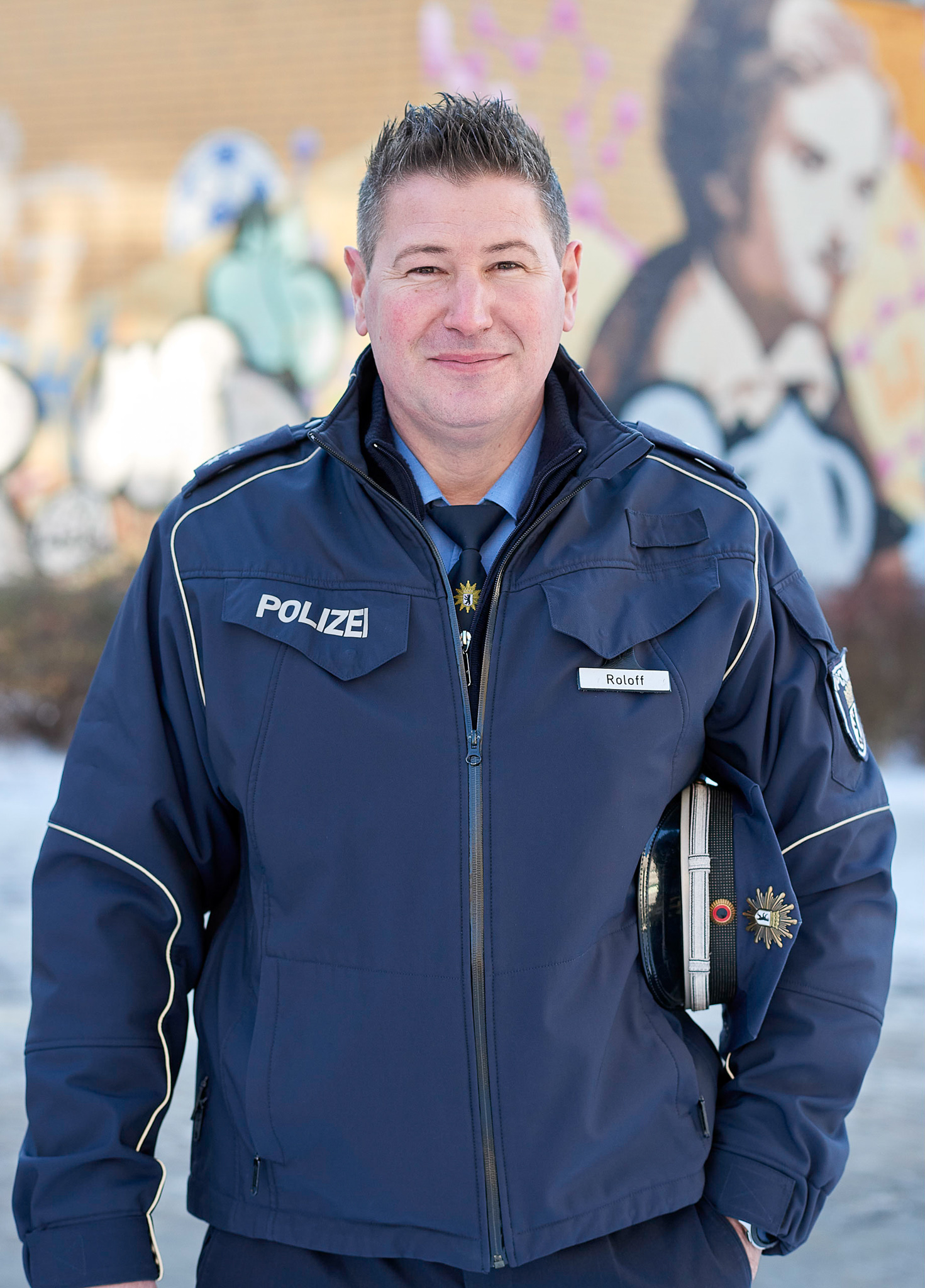 German police officer in a navy blue uniform standing outdoors in front of a colorful graffiti wall. The officer holds a police cap under one arm and wears a jacket with the word POLIZEI and a name tag reading Roloff. The environment is urban and the mood is professional and composed.
